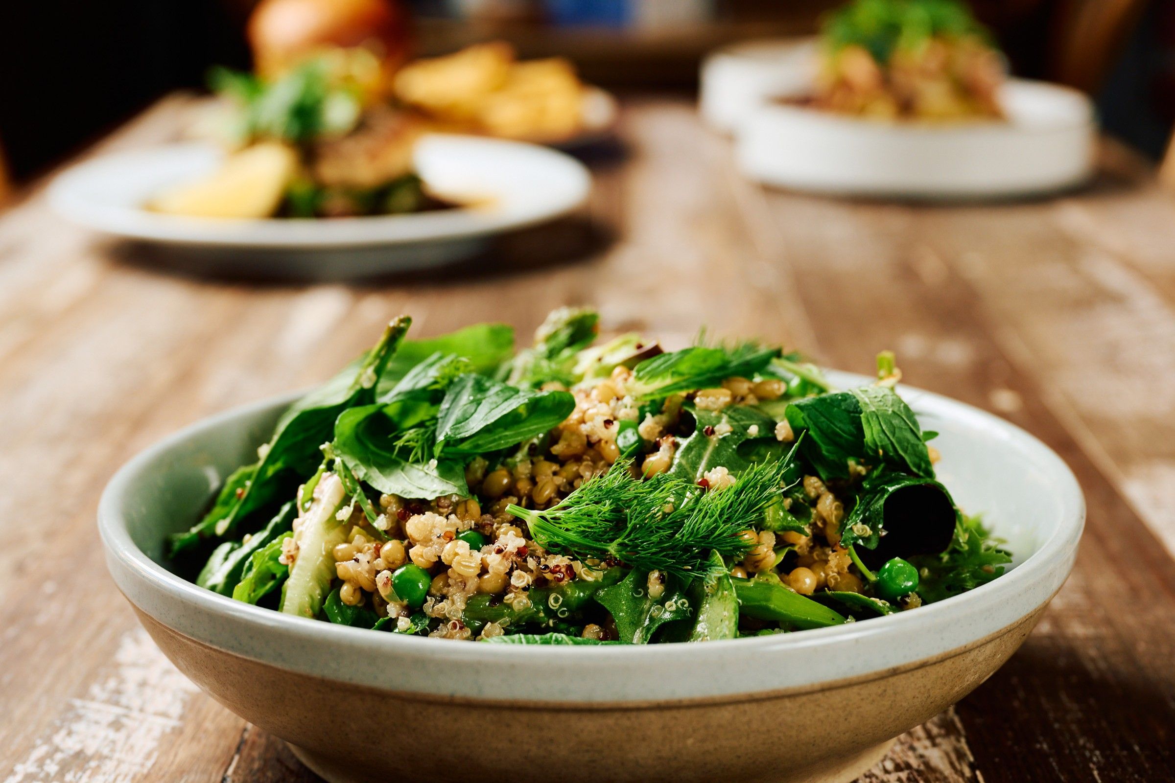A bowl of salad with various greens and grains, placed on a wooden table.