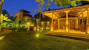 A well-lit outdoor area with a covered seating area, a stone building, and a pathway leading to a vehicle.
