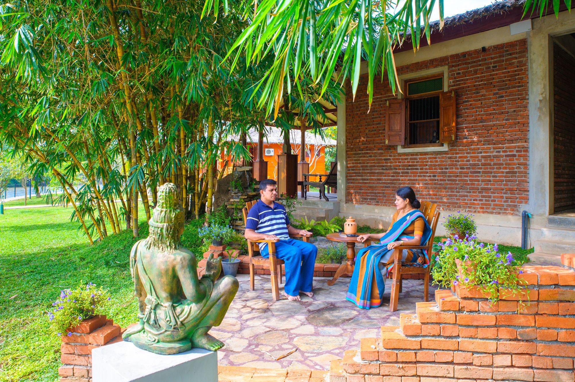 A courtyard with a statue, two people seated at a table, and a brick building in the background.