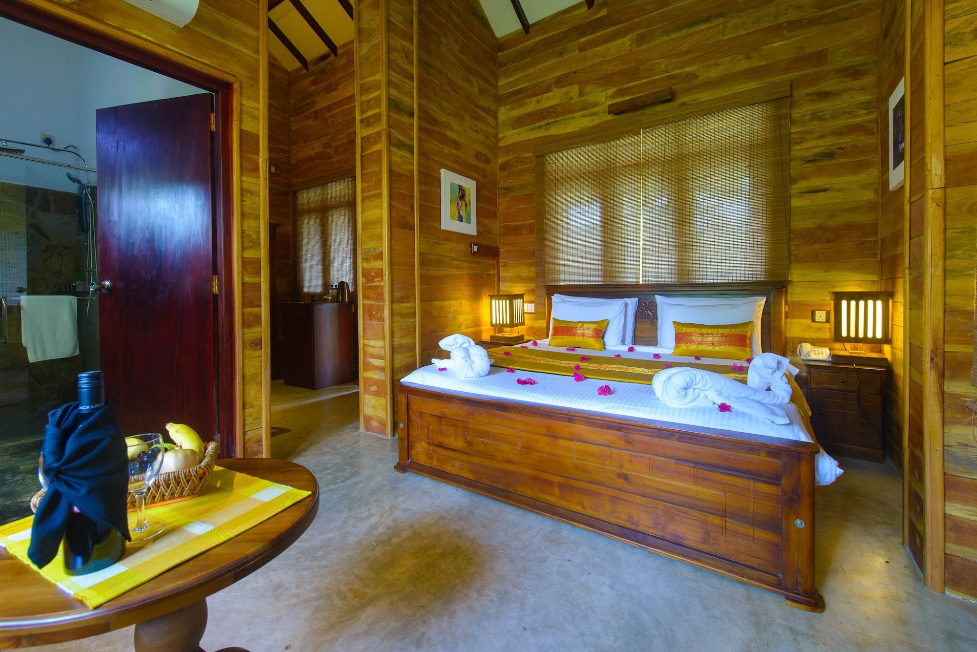 A wooden cabin-style bedroom with a large bed, a bedside table with a bottle and glass, and a wooden bench with a mat and fruit.