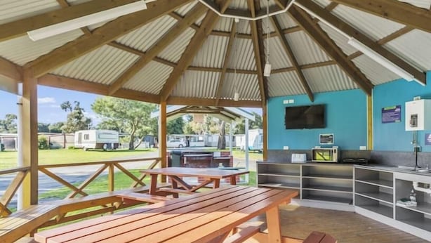 Un área de picnic techada con bancos de madera y una mesa, un televisor montado en la pared y vistas del parque circundante.