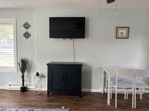 A room with a flat-screen TV mounted on the wall, a dark wooden cabinet, a white table with chairs, and a potted plant. - Perfect 1-bed house near downtown and Garden of the Gods Pet Friendly (Colorado Springs)