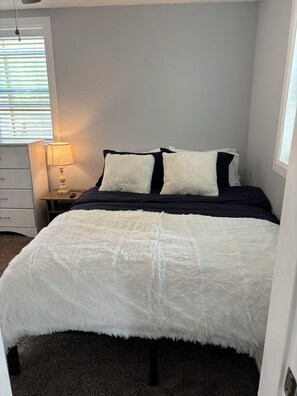 Un dormitorio con cama, mesita de noche con lámpara y ventana con persianas.