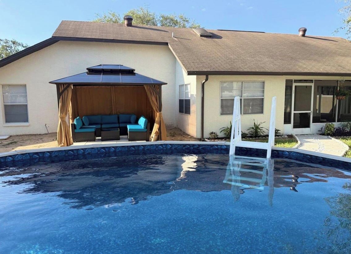 Una piscina residencial con un área de estar techada y una casa al fondo.