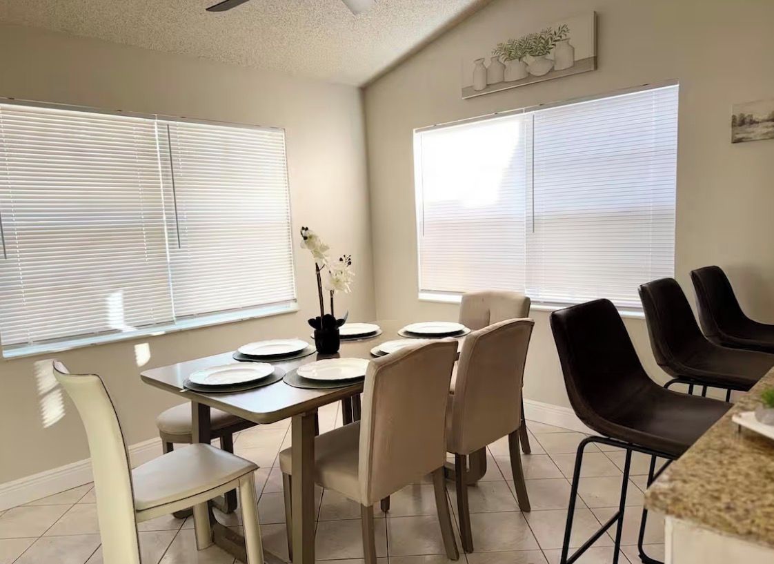Un comedor con una mesa dispuesta para cuatro personas, sillas alrededor y ventanas amplias con persianas.