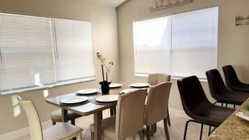 Un comedor con una mesa dispuesta para cuatro personas, sillas alrededor y ventanas amplias con persianas.