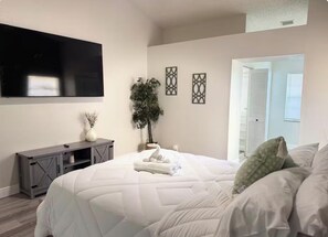 A bedroom with a large bed, a flat-screen TV mounted on the wall, a small cabinet, and a potted plant. - Spring Hill Stay 
Pool, Games & Relaxation (Spring Hill)