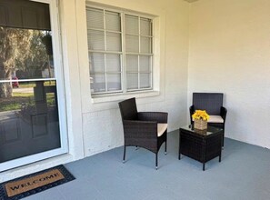 A porch with two wicker chairs, a small table with a vase of yellow flowers, and a window with blinds. - Spring Hill Stay 
Pool, Games & Relaxation (Spring Hill)