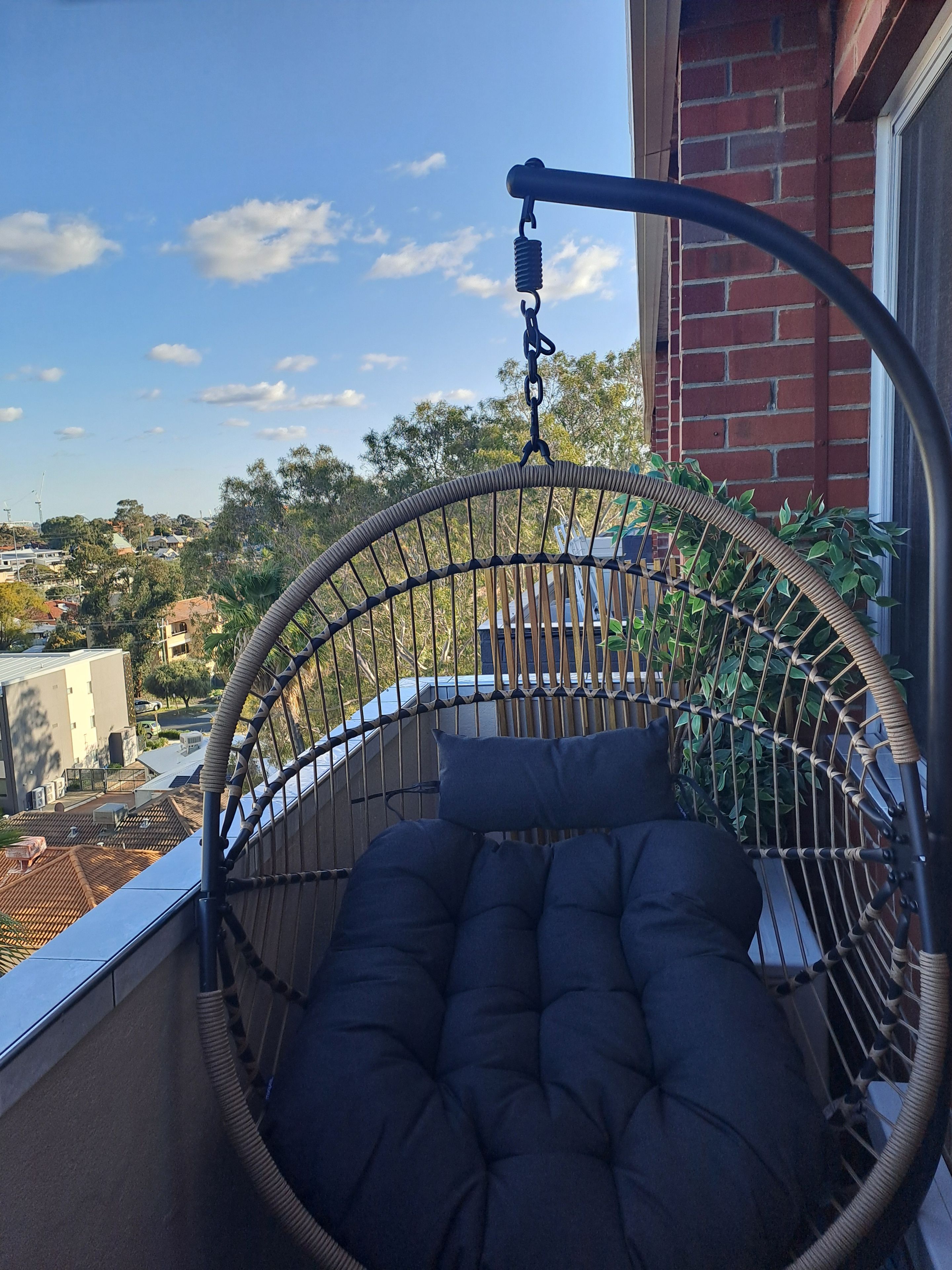 A hanging chair with a cushion, attached to a balcony railing, overlooking a residential area with houses and trees.