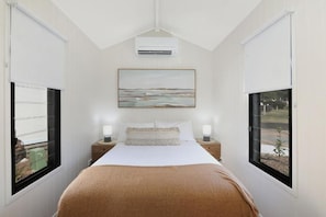 Quarto com cama, duas mesas de cabeceira, um quadro sobre a cama e duas janelas com persianas.
