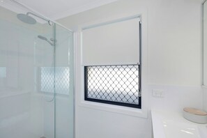 Casa de banho moderna com chuveiro de cabine de vidro, pequena janela com grade de metal e lavatório branco com bancada de madeira.