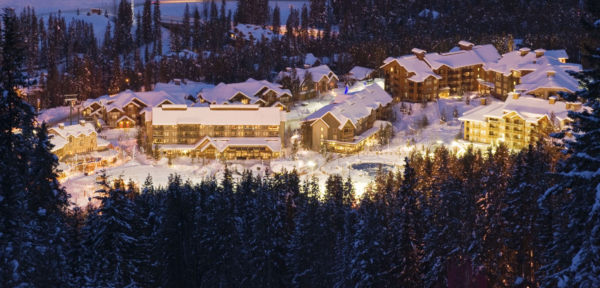 A snow-covered lodge with multiple buildings, including a large central structure and several smaller cabins, surrounded by dense forest.