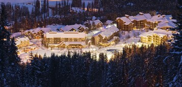 Un albergue cubierto de nieve con varias construcciones, incluyendo una estructura central grande y varias cabañas más pequeñas, rodeado de un bosque denso.