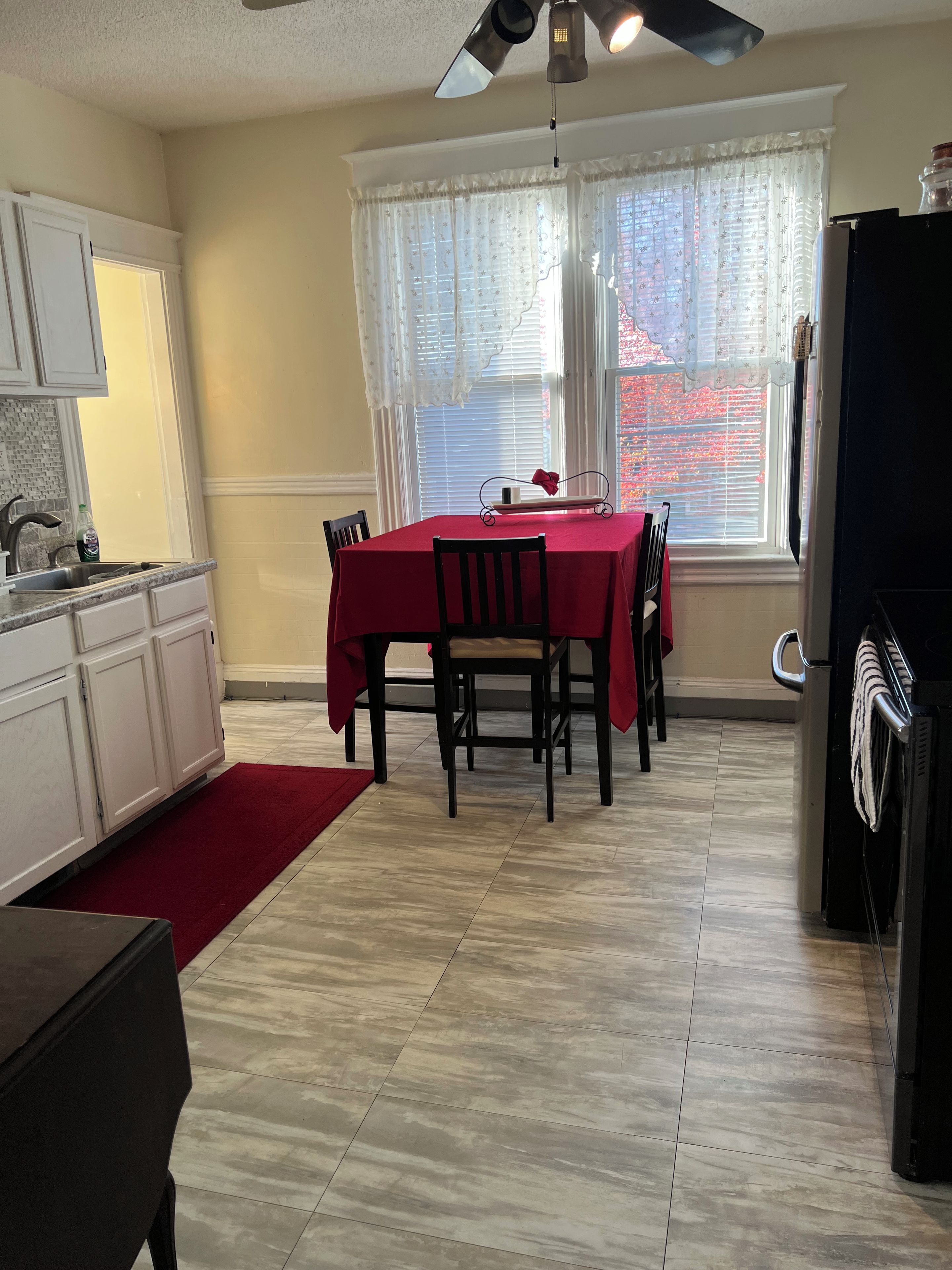 Uma área de refeições com mesa posta para quatro pessoas, cozinha com armários brancos, janela com persianas e ventilador de teto.