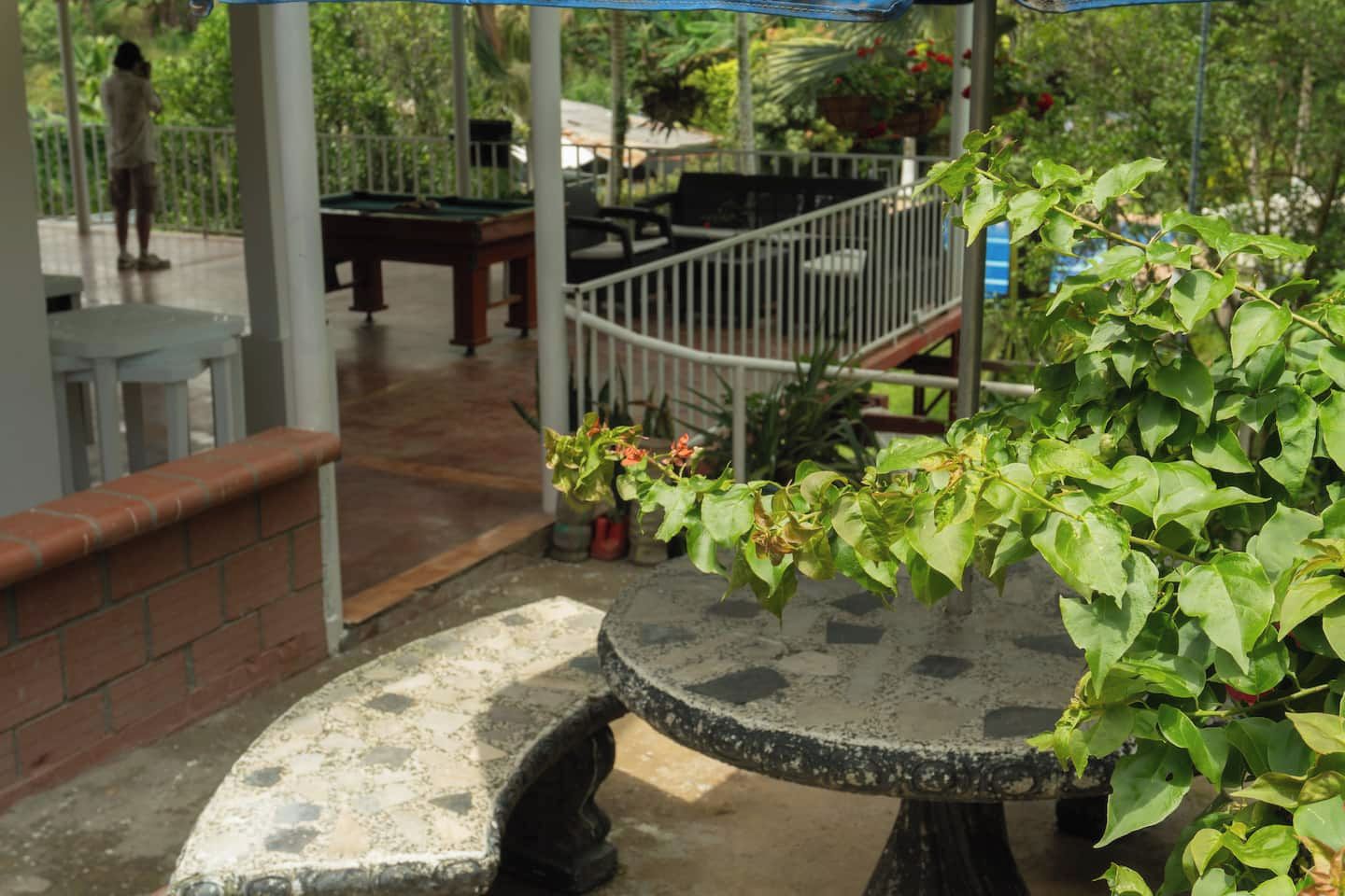 Un patio al aire libre con una mesa redonda y bancos, rodeado de follaje y una barandilla blanca.