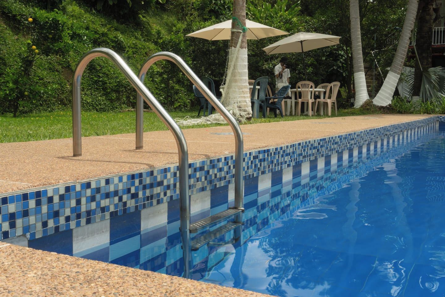 A swimming pool with metal steps leading into it, surrounded by a tiled edge and a grassy area with chairs and umbrellas.