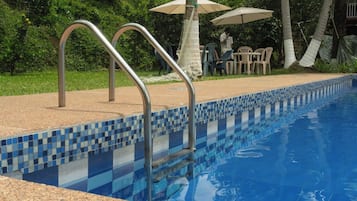 A swimming pool with metal steps leading into it, surrounded by a tiled edge and a grassy area with chairs and umbrellas.