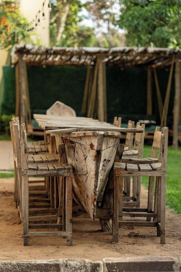 Un comedor al aire libre rústico con una larga mesa de madera y sillas, rodeado por una pérgola de madera.