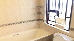 A bathroom with a bathtub, toilet, and a window with a towel hanging. - White House of Tanzania - Hostel (Arusha)