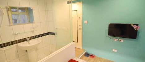 Un baño con lavamanos, espejo y ducha. Hay un televisor montado en la pared y una puerta que da a otra habitación.