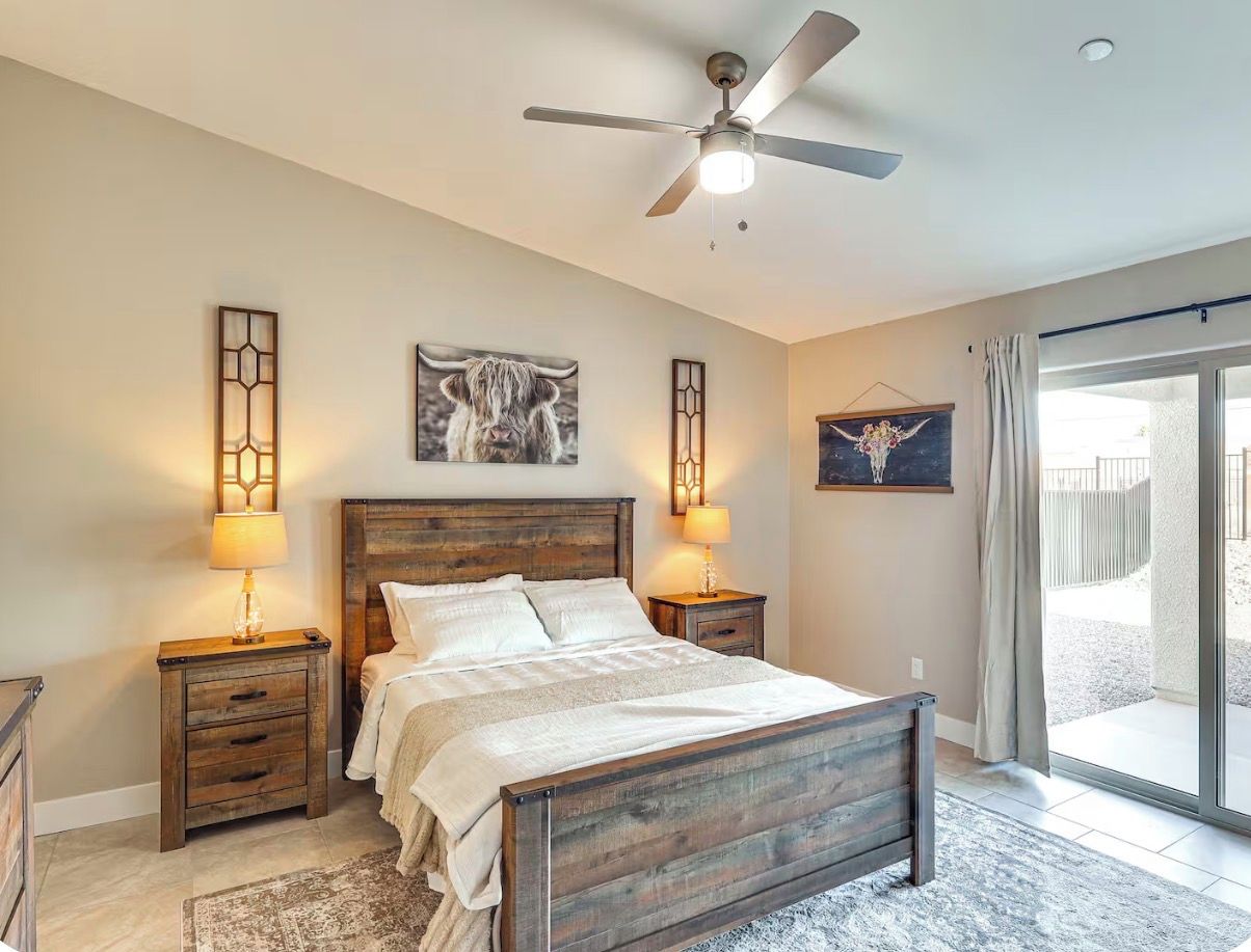 Une chambre à coucher avec un lit en bois, deux tables de chevet, un ventilateur de plafond et une vue sur l’extérieur, visible à travers une porte vitrée.