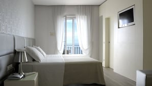 A hotel room with a bed, a bedside lamp, a TV mounted on the wall, and a view of the sky through sheer curtains. - Hotel Torremaura (Cervia)