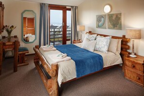A wooden-framed bed with a blue blanket, two bedside lamps, and a view of the sea through a large window. - Kowie River Chalets (Port Alfred)