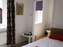 A bedroom with a bed, a small table with a cup and saucer, a window with a purple blind, and a framed picture on the wall.