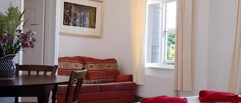 Habitación de hotel con cama, mesa de comedor, silla, sofá, ventana con cortinas e imagen enmarcada en la pared.
