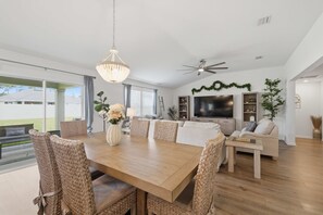 Un vaste espace de vie comprenant une table à manger en bois, des chaises en osier, un lustre, un ventilateur de plafond, un téléviseur à écran plat et une grande fenêtre donnant sur l’extérieur.