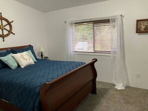 A bedroom with a bed, a window with blinds, a wall-mounted ship's wheel, and a small bedside table with a lamp.
