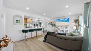 A modern living room with a sofa, a dining area with bar stools, a kitchen with a refrigerator, and a TV mounted on the wall.