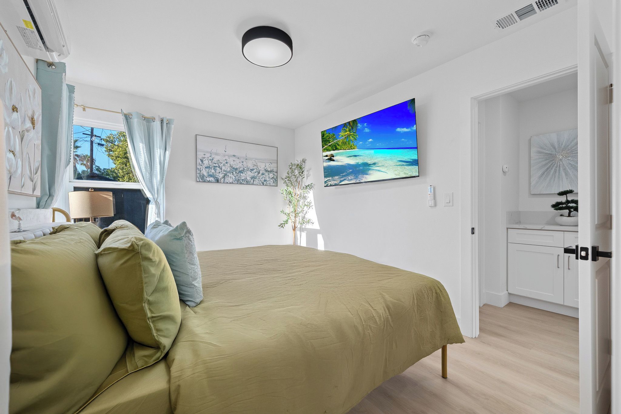 A bedroom with a bed, a window with curtains, a wall-mounted TV, and a door leading to a bathroom.