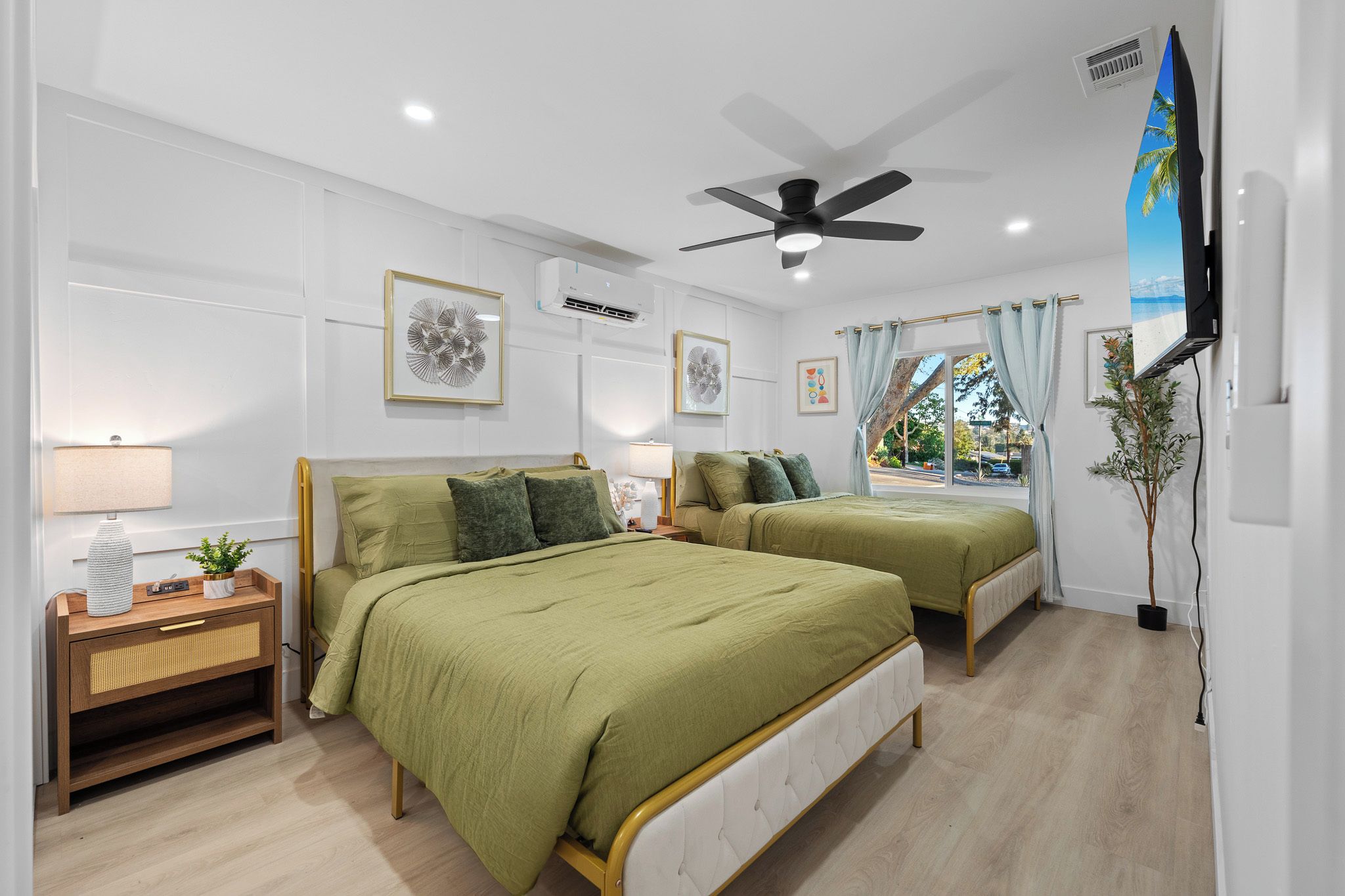 A hotel room with two beds, a ceiling fan, artwork on the walls, and a window with curtains.