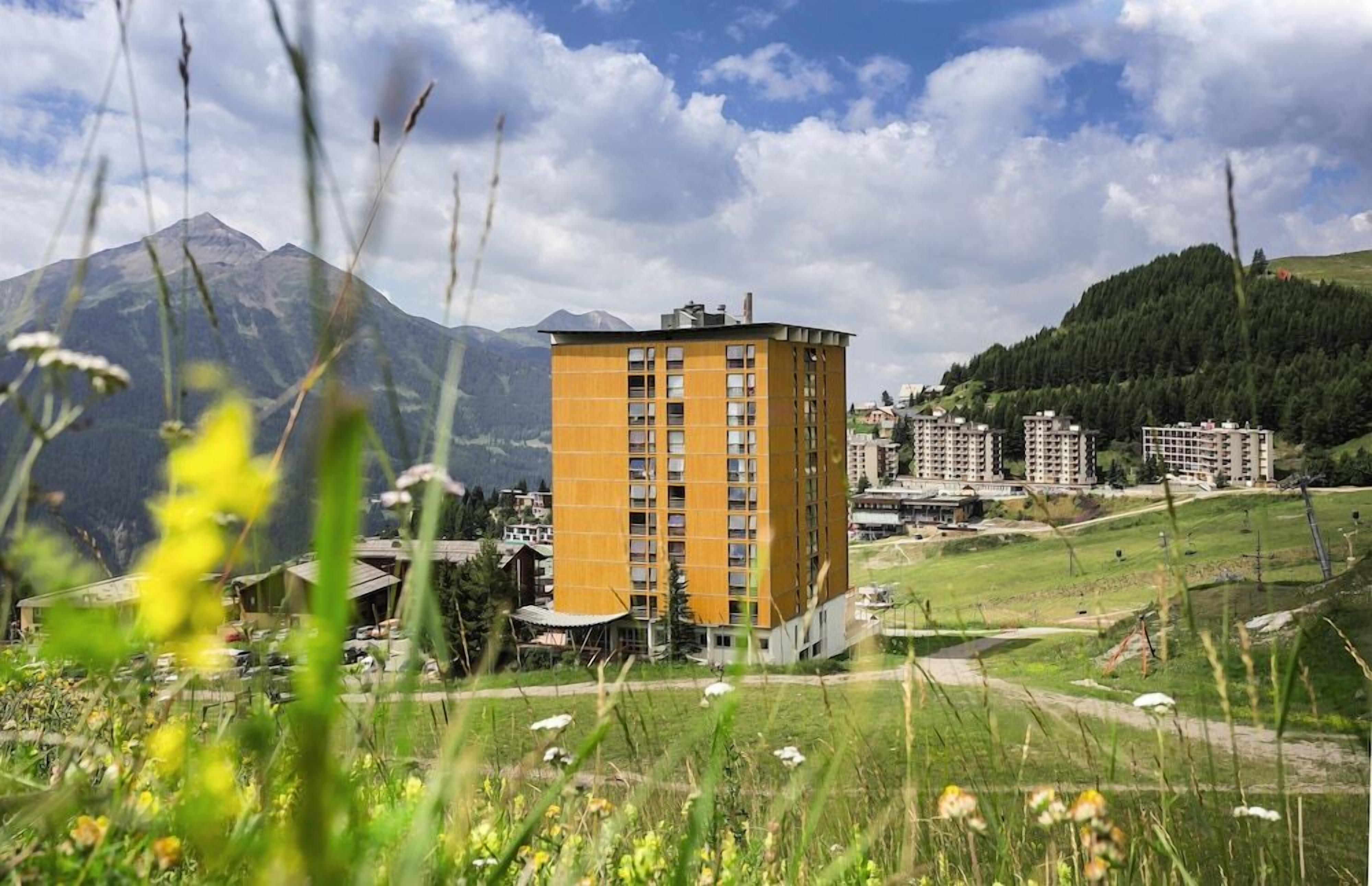 A multi-story building with a flat roof, surrounded by a grassy area and mountains in the background.