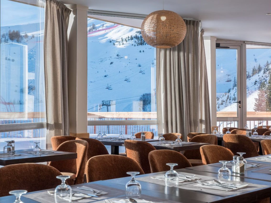A restaurant with tables set for dining, large windows with curtains, and a view of a snowy mountain area.