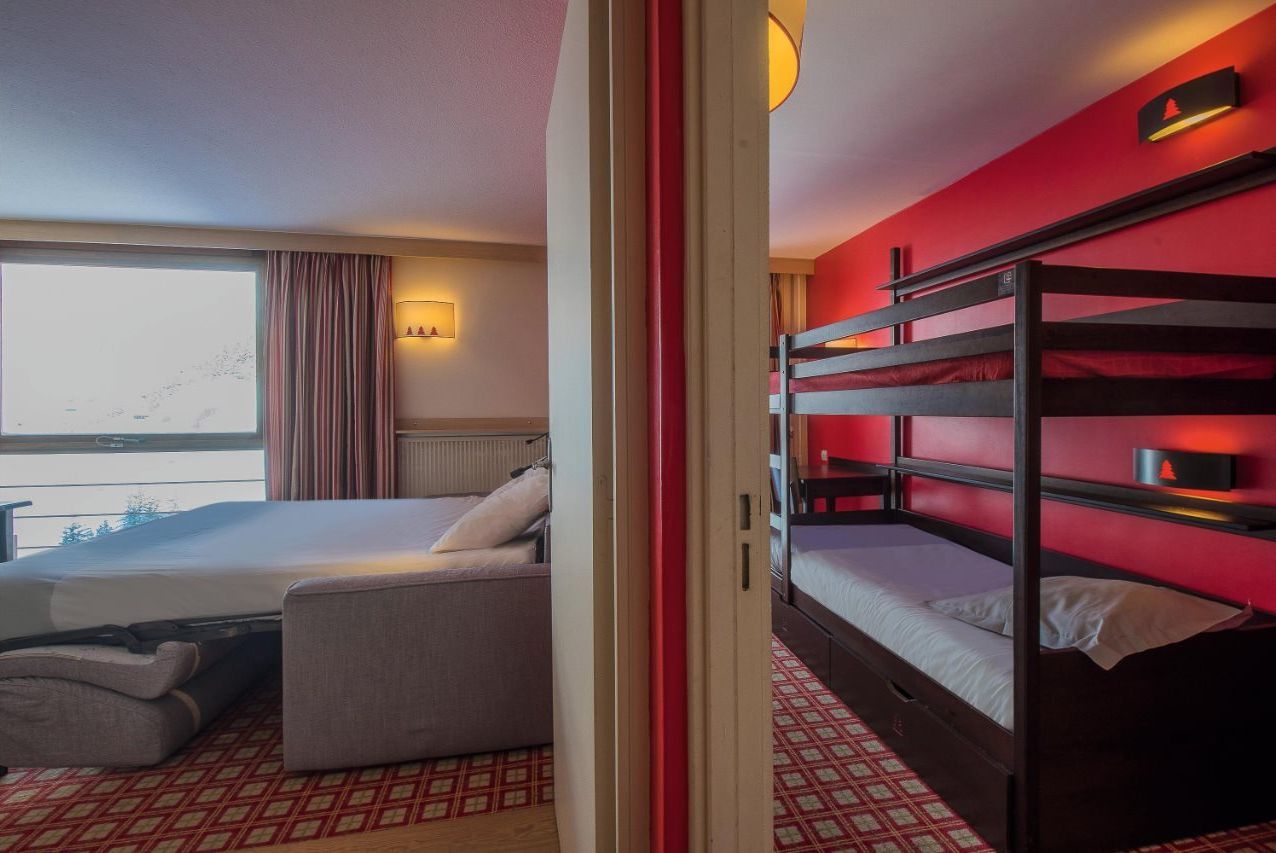 A hotel room with a bunk bed, a single bed, a sofa, a window with curtains, and a wall-mounted lamp.