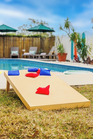 Ein Poolbereich mit einer Holzliege, einem kleinen Tisch mit Kissen und einem Pool im Hintergrund.