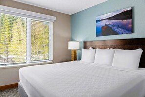 Une chambre d’hôtel avec un grand lit, une fenêtre avec des stores, une table de chevet, une lampe et un tableau au mur.