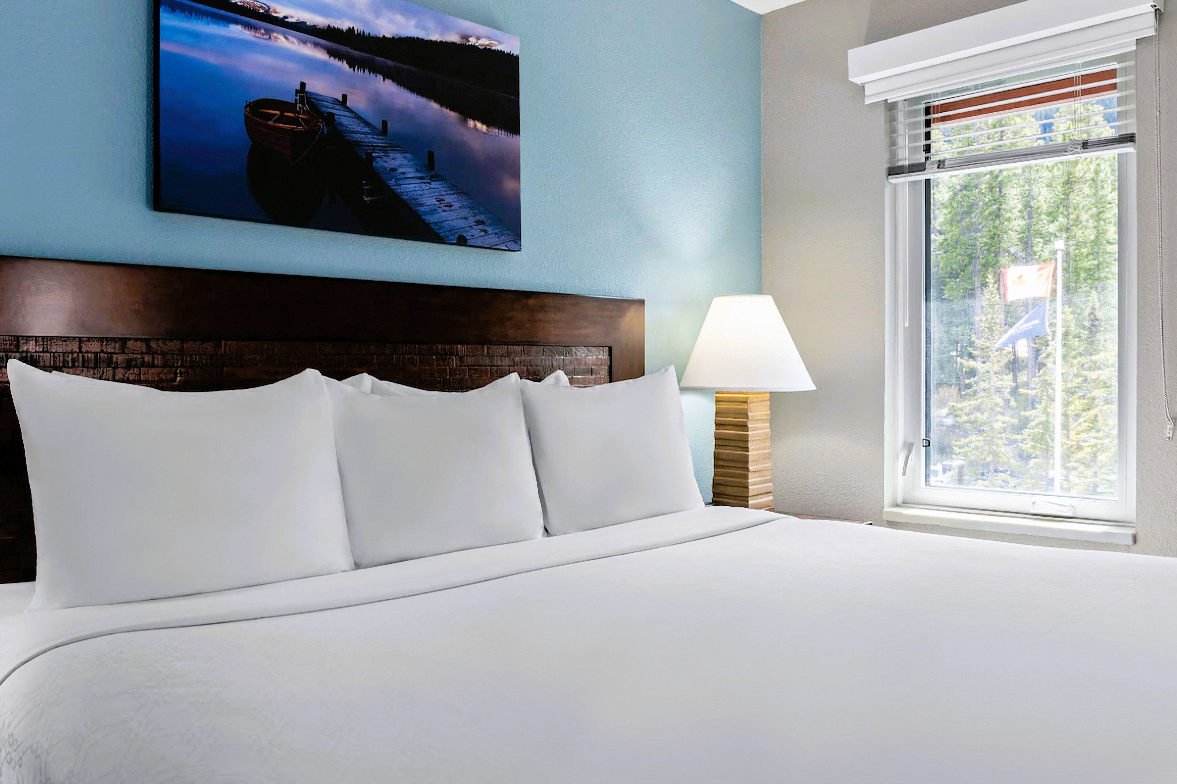 A hotel room with a large bed, a headboard with a brick pattern, a wall-mounted picture of a lake, a window with blinds, and a bedside lamp.