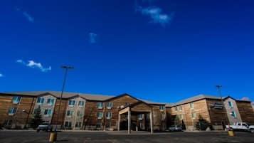 A multi-story hotel with a central entrance, parking lot, and a clear blue sky.