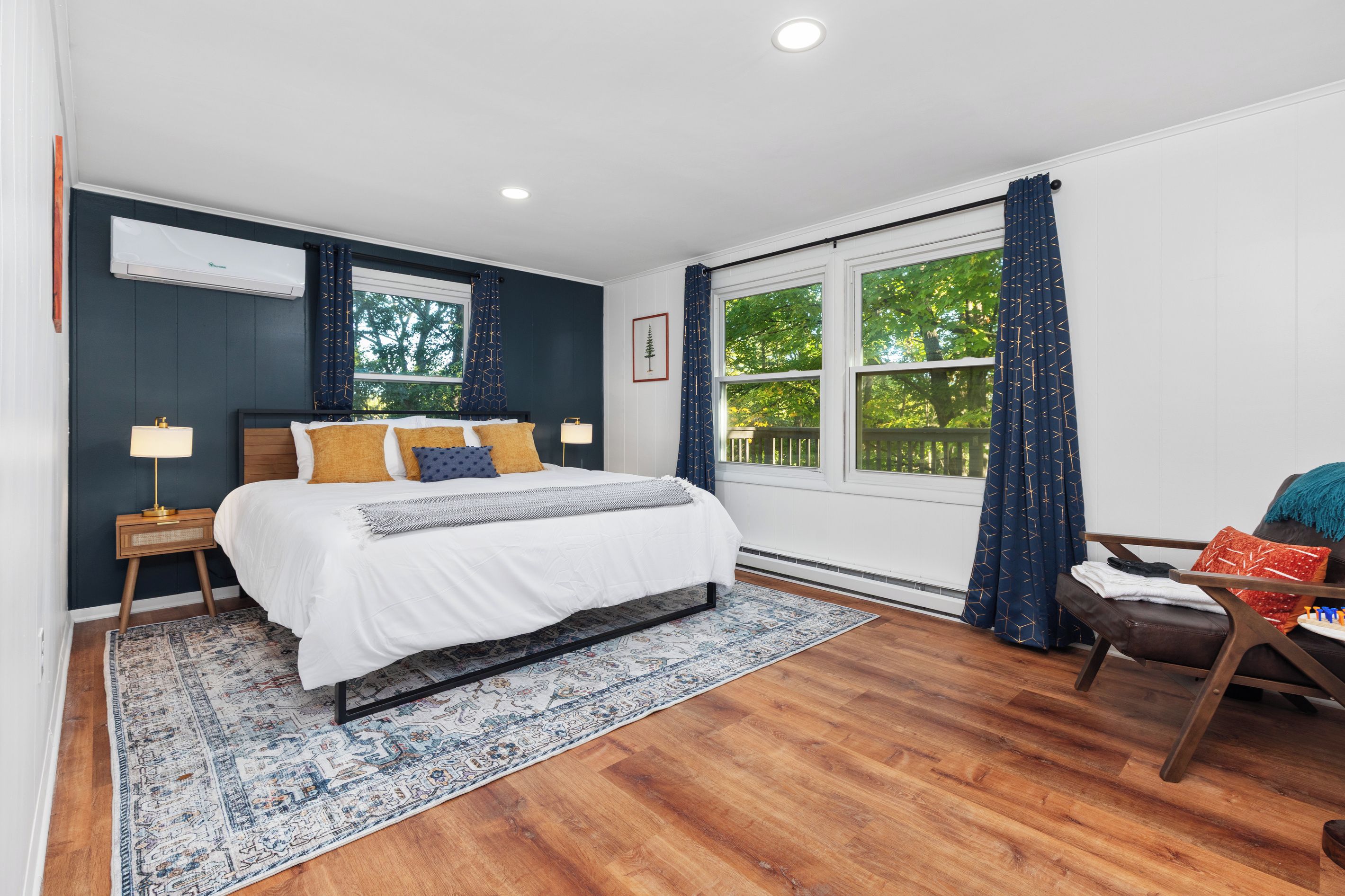 Une chambre à coucher avec un lit, des tables de chevet, une fenêtre avec des rideaux, une vue sur les arbres et un plancher en bois.