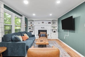 A living room with a sofa, coffee table, ottoman, fireplace, television, and a large window with curtains. - Cottage Nest at Downtown Berkeley Springs (Berkeley Springs)