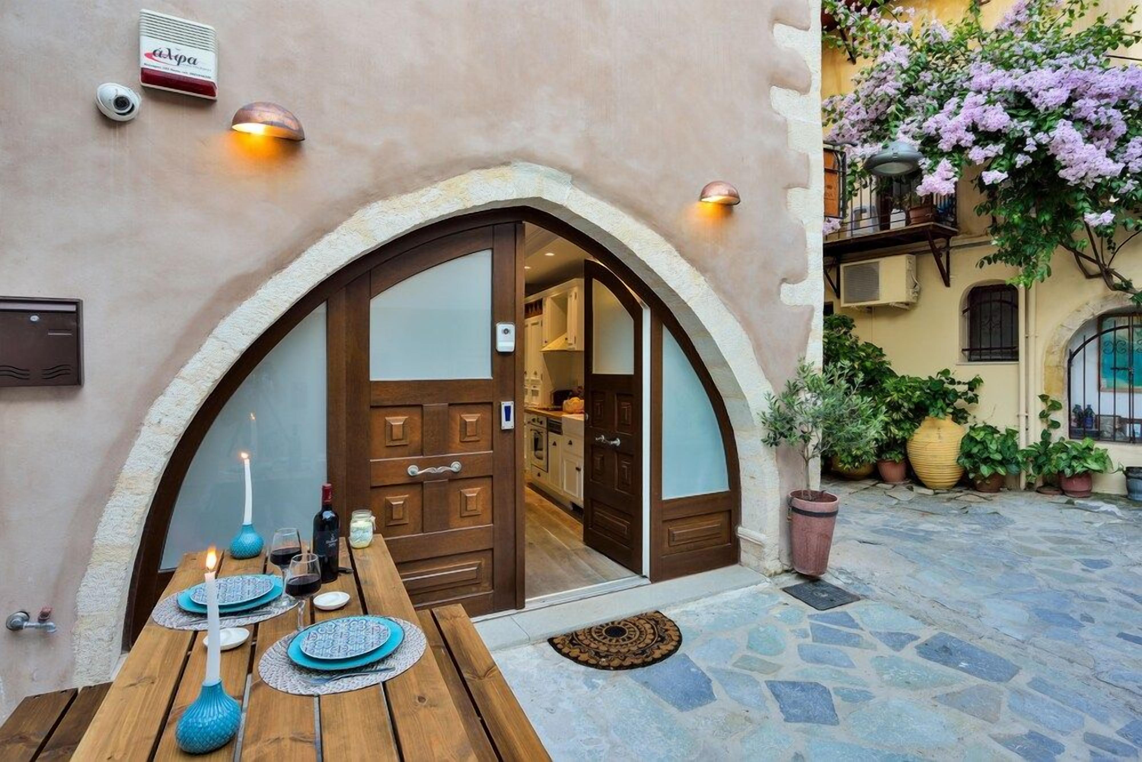 An outdoor dining area with a wooden table set for two, a door leading to a room, and potted plants.