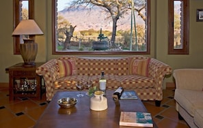 A cozy living room with a checkered sofa, a coffee table with a book, a lamp, and a view of a mountainous landscape through a large window.