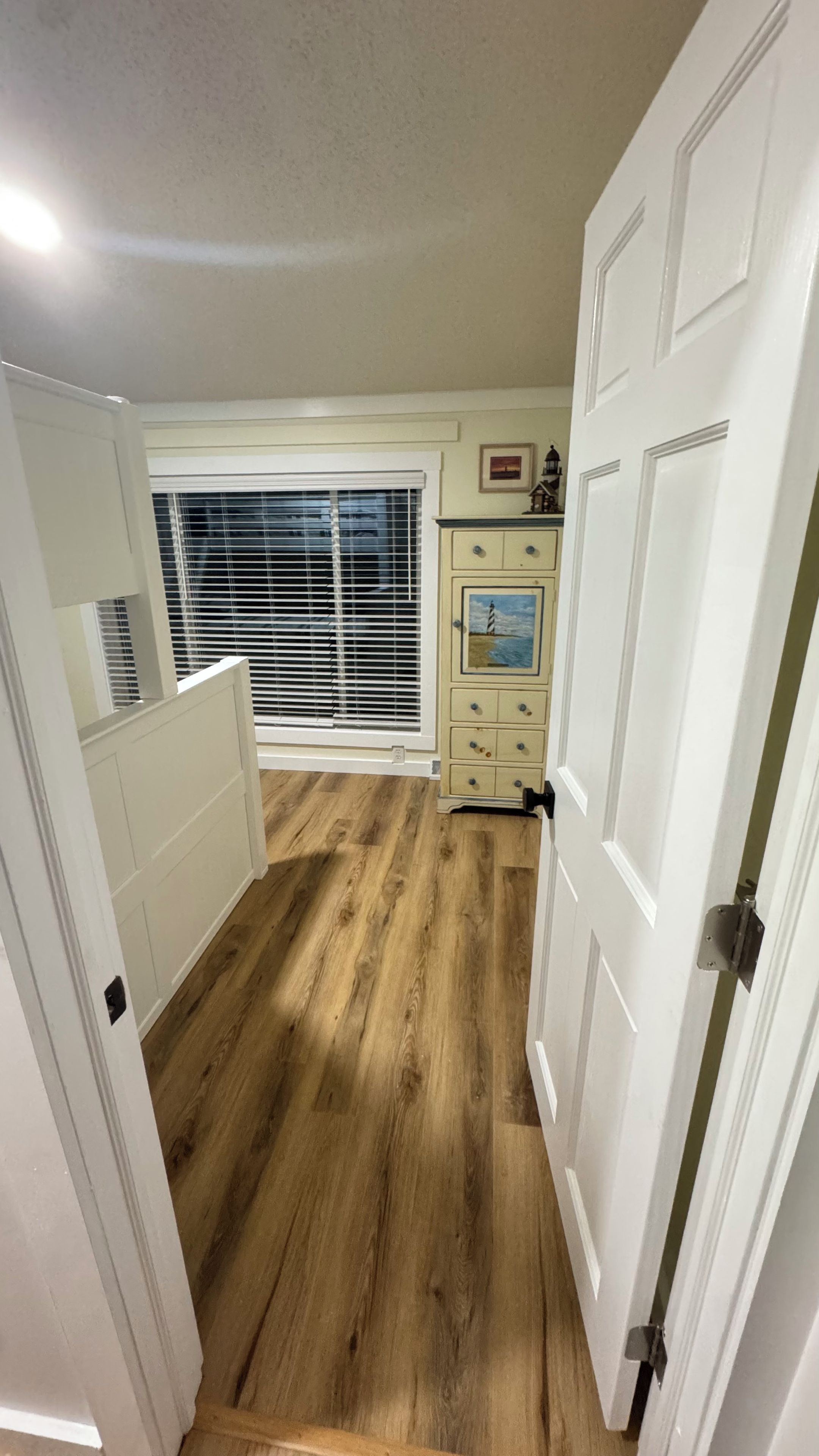 A narrow hallway with wooden flooring, white walls, and a window with blinds.