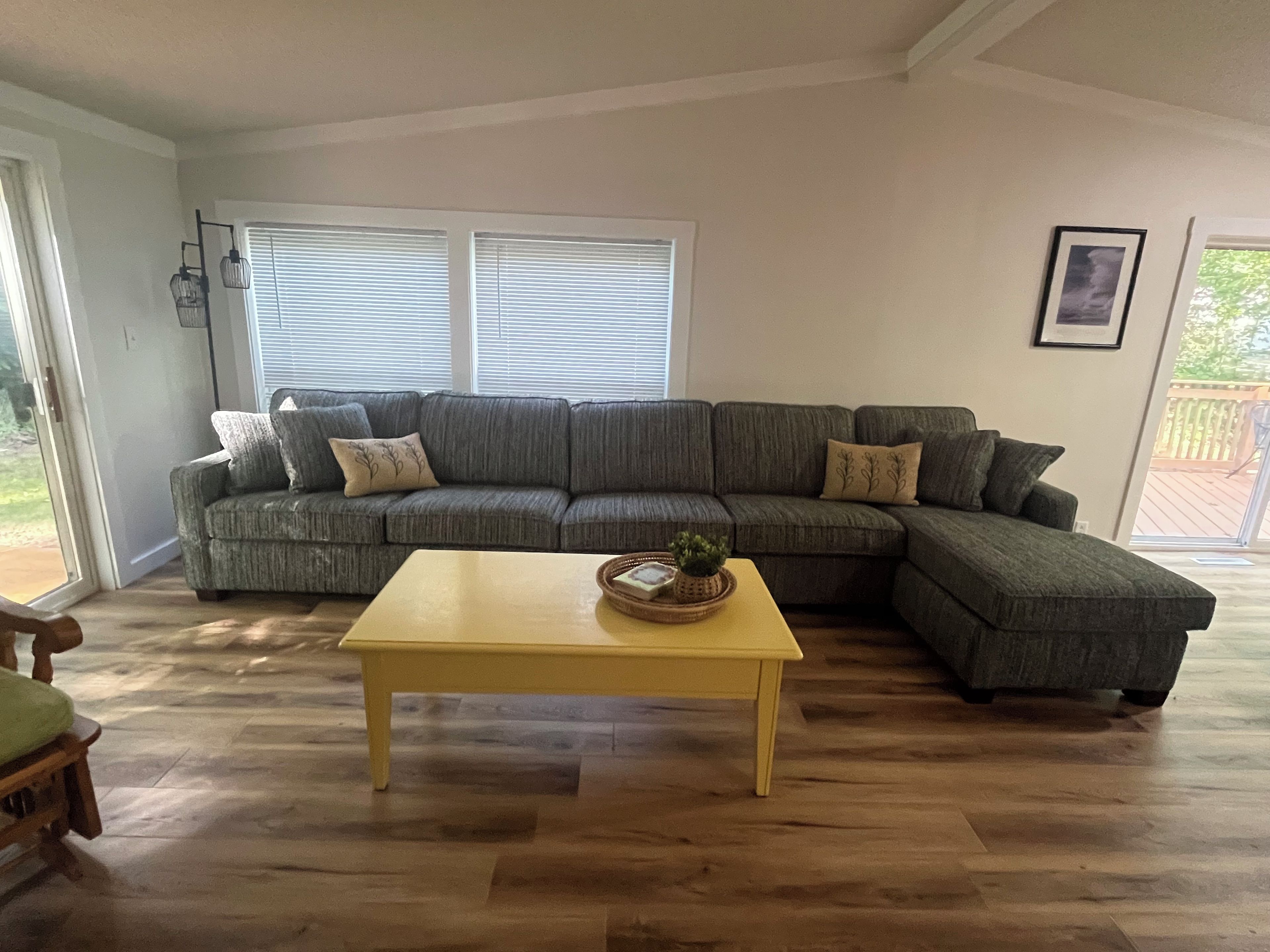 Un salon avec un canapé d’angle, une table basse jaune et une grande fenêtre équipée de stores.