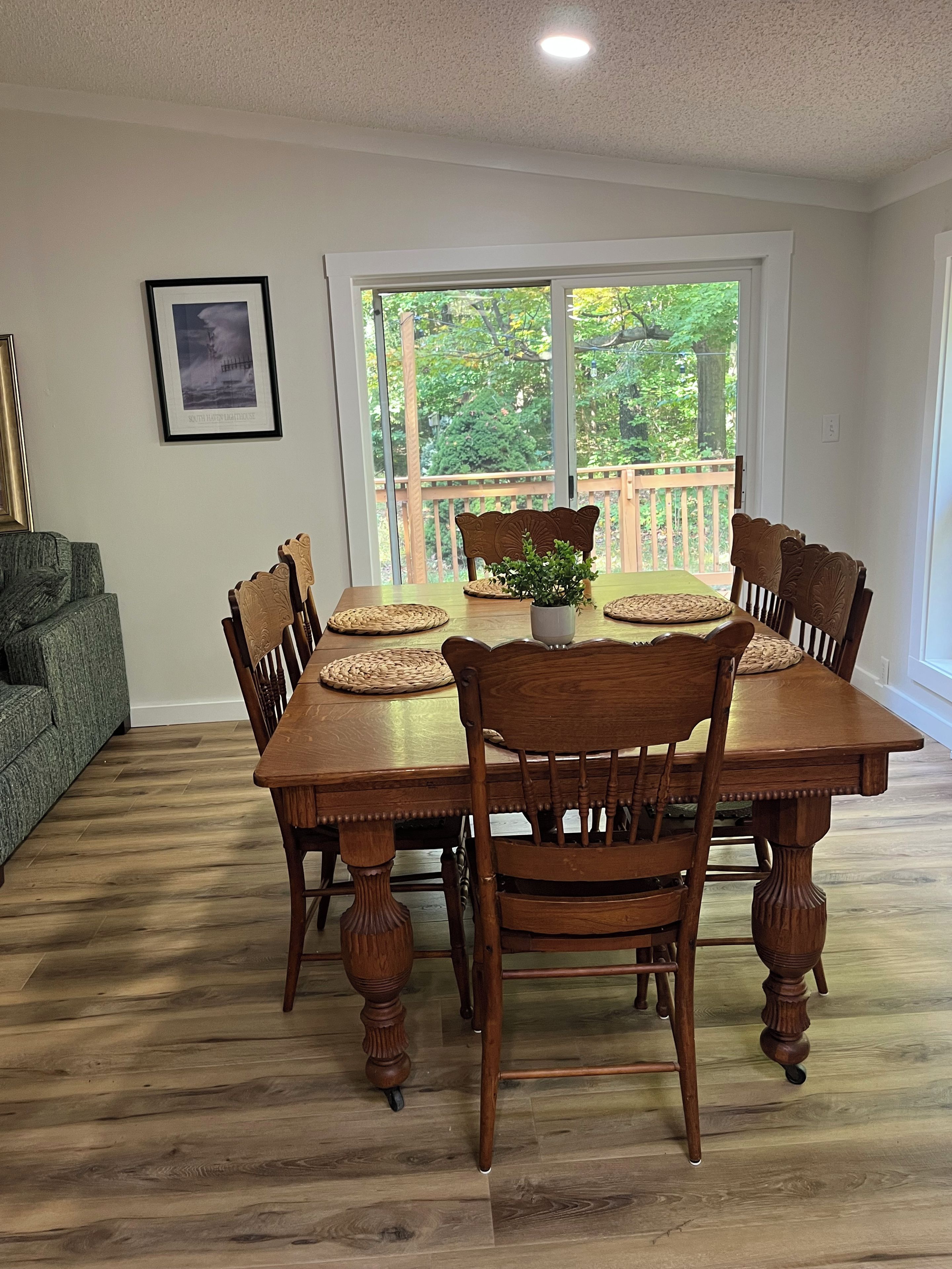 A dining room with a wooden table and chairs, a potted plant, and a view of a balcony with greenery.