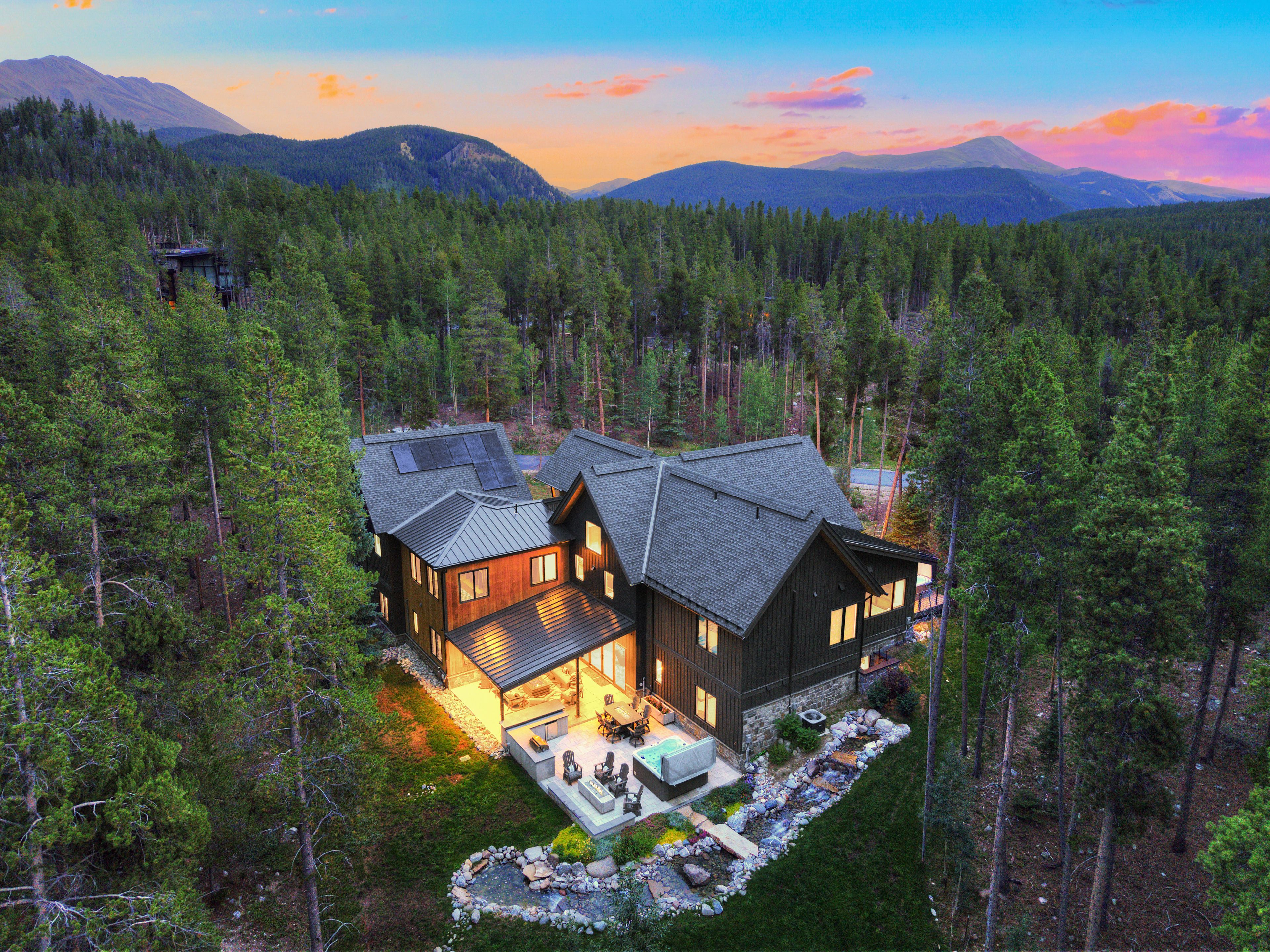 A modern cabin with a large deck, surrounded by dense forest and mountains in the background.