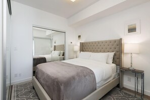 Quarto de hotel com uma cama grande, mesa de cabeceira com abajur, espelho e uma pequena imagem na parede.